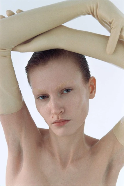Close-up portrait of fair-skinned adult woman with neutral expression, wet-look hair, and wearing long latex gloves against white background.