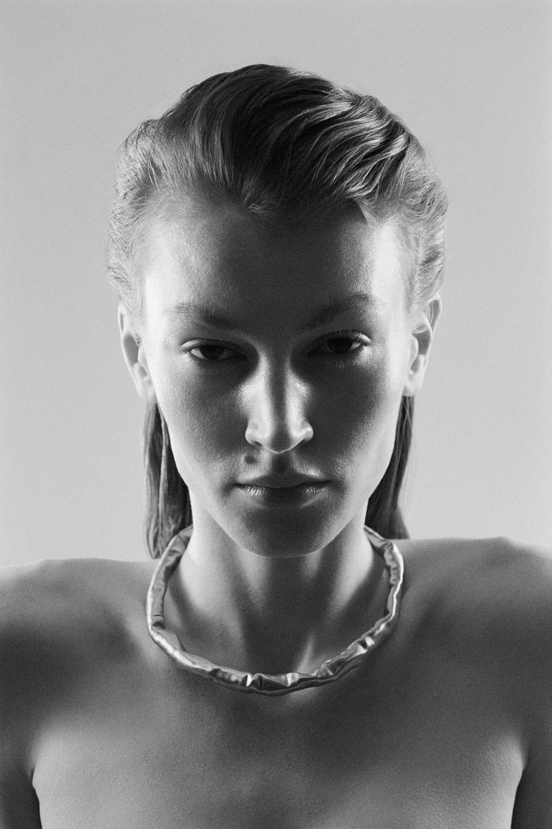 Striking close-up studio portrait of a fair-skinned woman with slicked-back hair, wearing a unique silver necklace, set against a plain background.