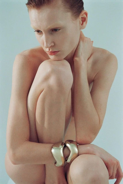 Close-up portrait of a fair-skinned person with wet red hair and simple gold bracelets in a studio with a light blue background.