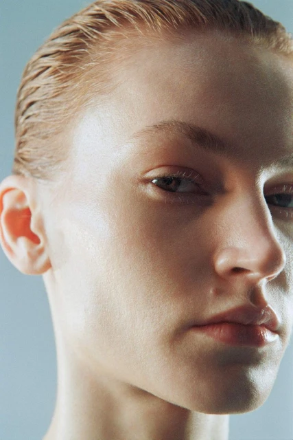 Close-up portrait of a woman with slicked-back wet-look auburn hair, fair skin, gazing at the camera with a serious expression in diffused lighting.