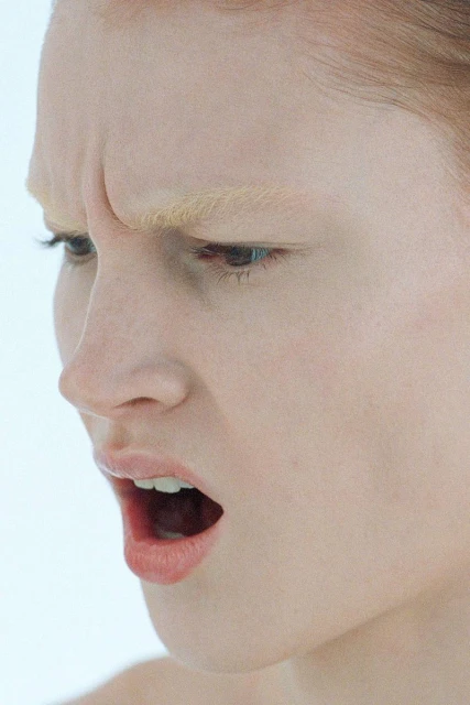 Extreme close-up of a fair-skinned woman's face expressing distress, mouth open, against a stark white backdrop in a studio setting.