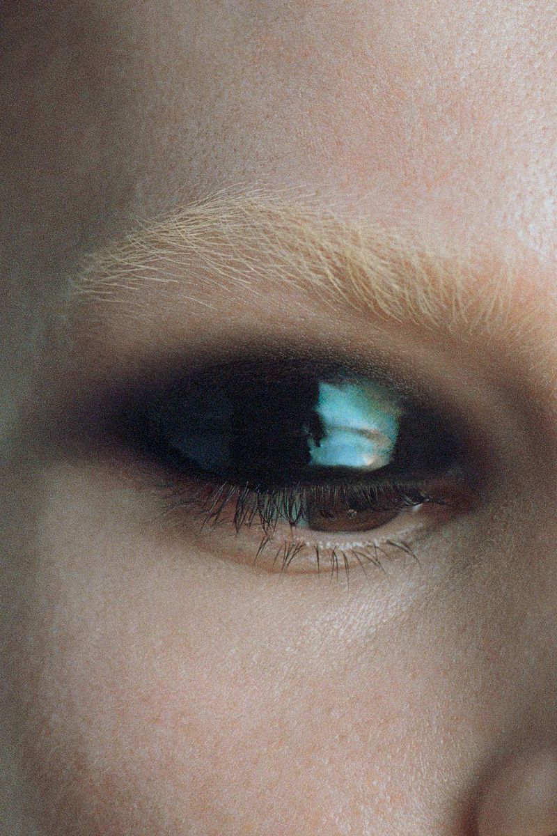 Macro shot of fair-skinned adult female face focusing on a dark brown eye, blonde eyebrow, and skin texture in an indoor setting.