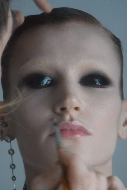 Close-up of a light-skinned adult woman with black eyeshadow, glossy pink lips, and light eyebrows having makeup applied by multiple hands.