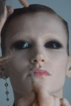 Close-up of a light-skinned adult woman with black eyeshadow, glossy pink lips, and light eyebrows having makeup applied by multiple hands.