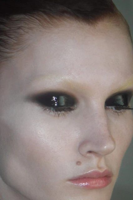 Close-up studio shot of a fair-skinned adult female with dark smoky eye makeup, glossed lips, and slicked-back hair. Beauty advertising.