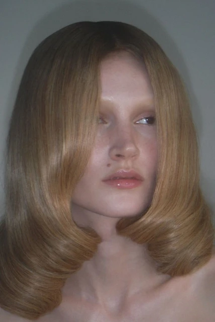 Close-up studio shot of a woman with blonde wavy shoulder-length hair, fair skin, and glossy lips, gazing off-camera with neutral expression.