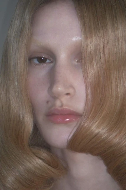 Close-up portrait of a young adult woman with long blond hair partially obscuring her face. Her gaze is direct, with light skin tone.