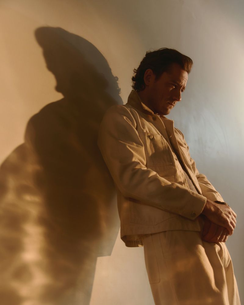 Man in tan jacket leans against a white wall, creating a shadow effect. Portrait shot, neutral expression, studio lighting.