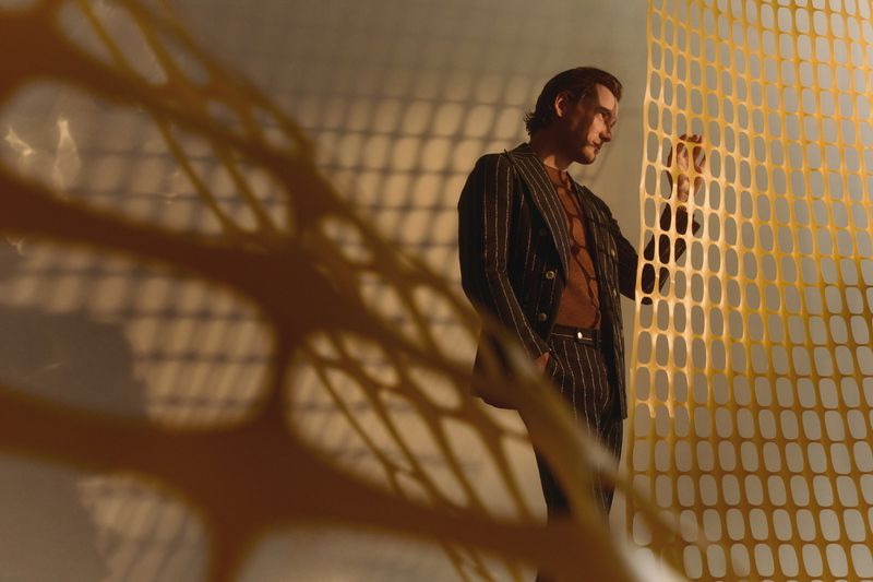 Adult male in a striped suit stands near a yellow net in a studio with soft, directional lighting creating patterns on the wall behind.