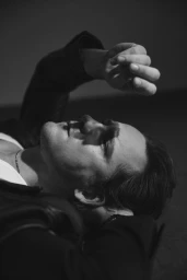 Portrait of actor Lewis Pullman lying down with his arm raised, wearing a jacket, captured in dramatic black and white monochrome.