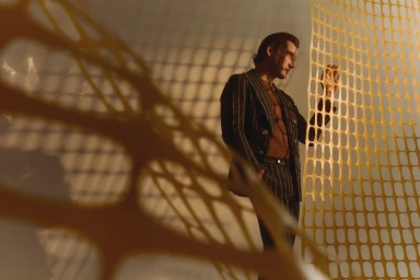 Actor Lewis Pullman wearing a pinstripe suit standing behind an orange plastic mesh barrier casting patterned shadows.
