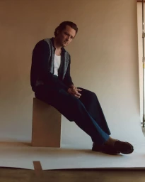 Actor Lewis Pullman seated on a wooden block wearing a dark patterned cardigan, white tank top, and trousers against a seamless backdrop