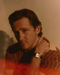 Portrait of Lewis Pullman wearing a patterned brown polo shirt and a wristwatch, posed against a warm wall.