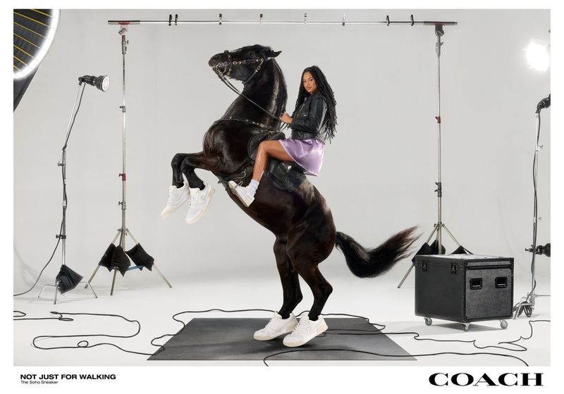 A woman in sneakers rides a rearing black horse in a photo studio, modeling for Coach, with lighting equipment and a black platform.