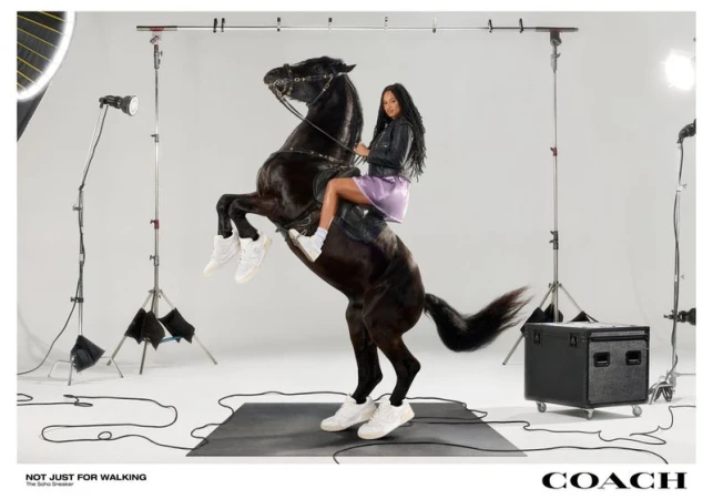A woman in sneakers rides a rearing black horse in a photo studio, modeling for Coach, with lighting equipment and a black platform.