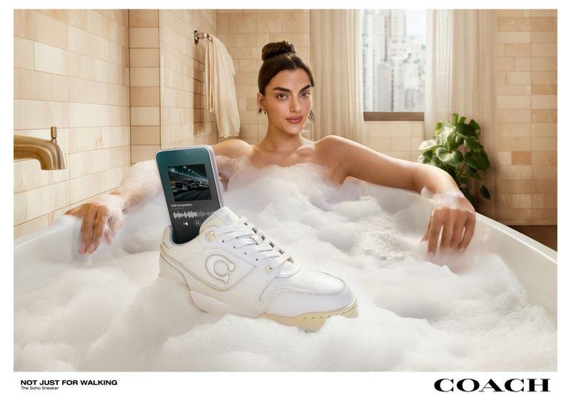 Woman in a bathtub with bubbles, a phone, and a white Coach sneaker in the foreground, against a neutral bathroom backdrop.