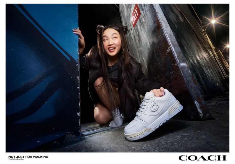 A young Asian female model poses in a gritty urban setting, holding a white Coach sneaker as part of a fashion editorial shoot.