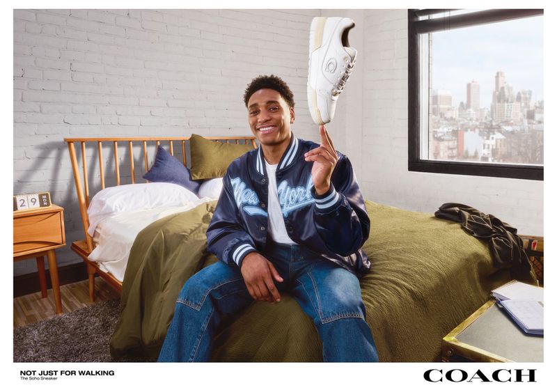 A young man sits on a bed balancing a white sneaker on his finger, indoors, with a cityscape view from a window in the background.