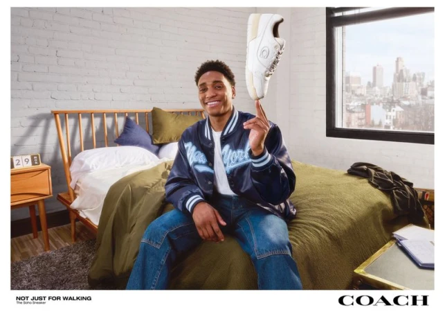 A young man sits on a bed balancing a white sneaker on his finger, indoors, with a cityscape view from a window in the background.