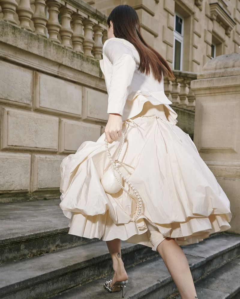 Woman in a light-colored, layered dress walks up stone steps outdoors, carrying a small purse, with a city building in the background.