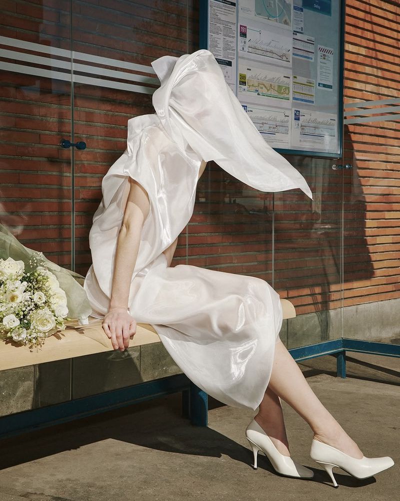 Woman in flowing white fabric sits on bus stop bench with floral bouquet, brick and glass background, soft light, fashion and style.