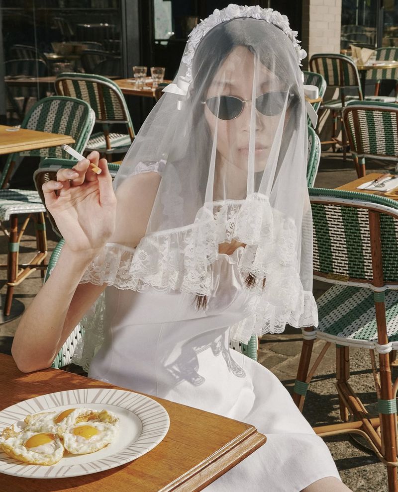 Woman in white dress and veil sits at an outdoor cafe, smoking cigarette with plate of fried eggs, surrounded by chairs.