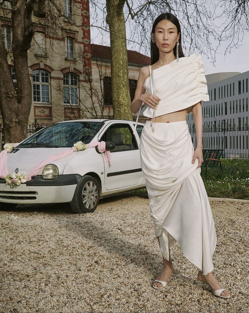Elegant Asian woman in a white draped outfit poses next to a decorated white compact car with city buildings and trees in the background.