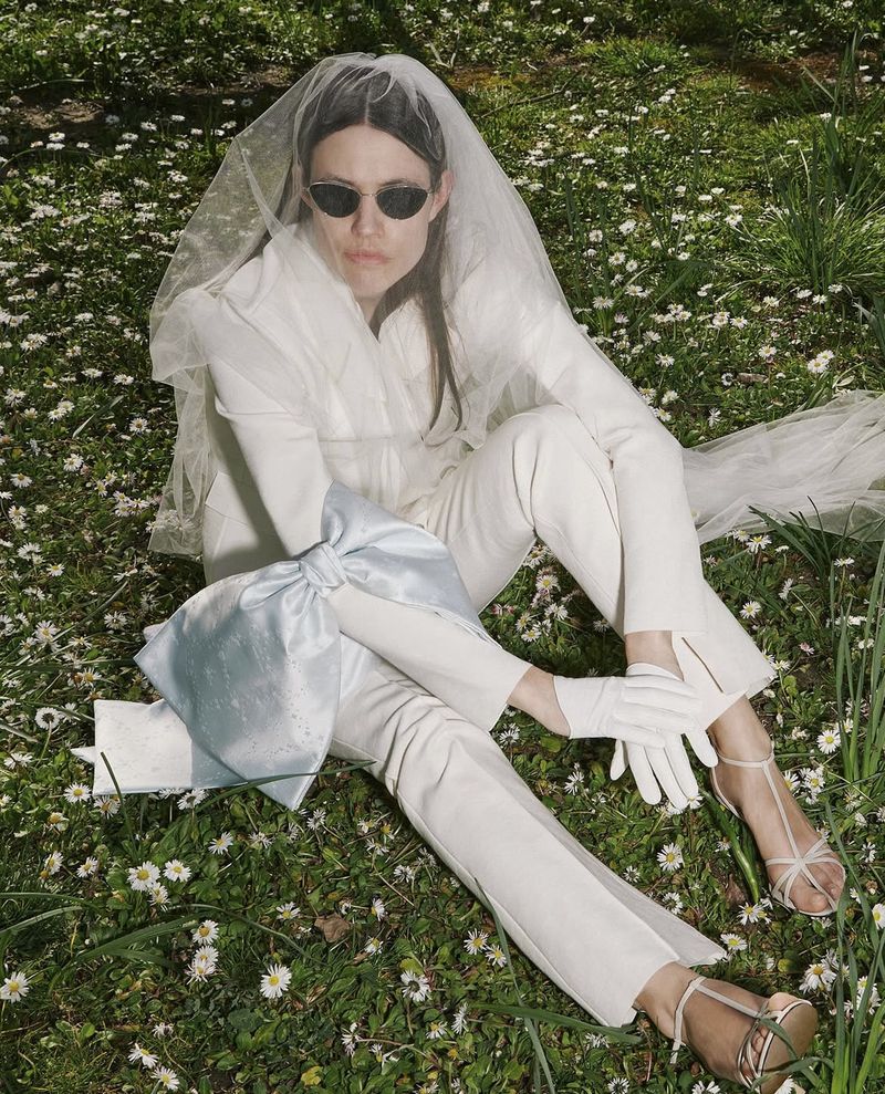 Woman in a white suit and veil sits in a field of daisies holding a blue bow bag, complemented by white gloves and strappy sandals.