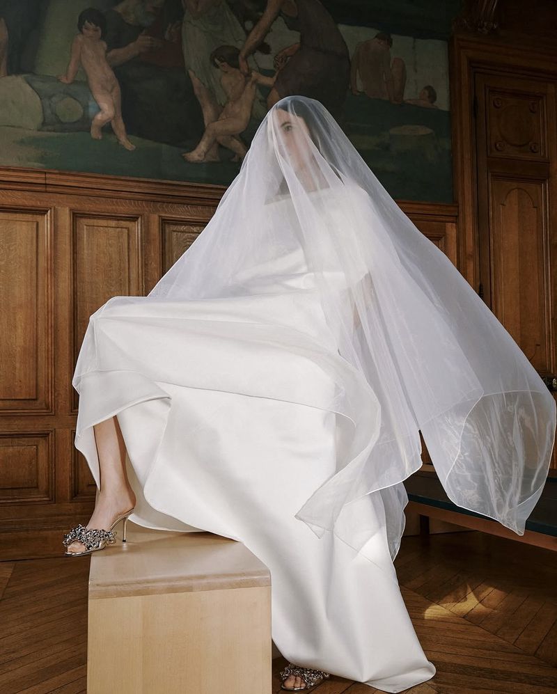 Woman in a white wedding dress and veil poses on a wooden box in an indoor space with wood paneling and a mural in the background.