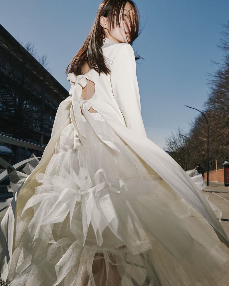 Elegant adult female in a white dress posing on a city street with trees and a blue sky in the background, fashion editorial.
