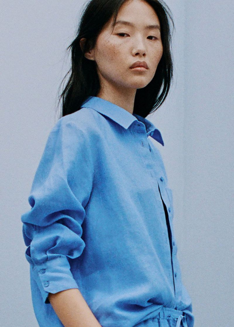 Portrait of a young adult woman with long dark hair wearing a button-down cornflower blue shirt in a naturally lit photo studio.