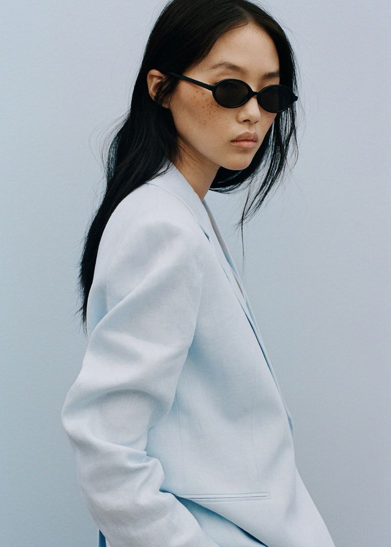 An Asian woman poses in a light blue blazer and black sunglasses against a solid light blue background in a fashion or beauty context.