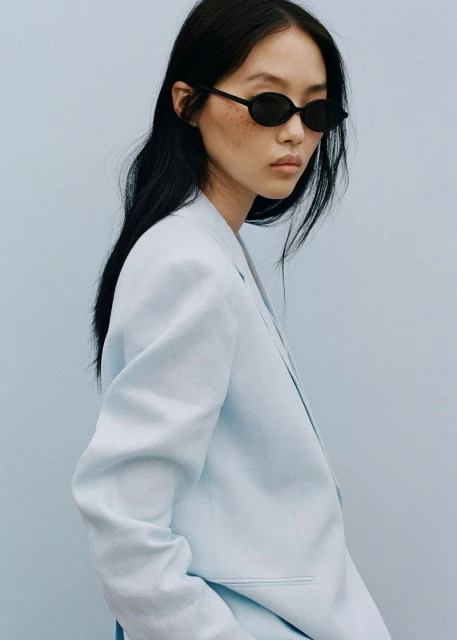 An Asian woman poses in a light blue blazer and black sunglasses against a solid light blue background in a fashion or beauty context.