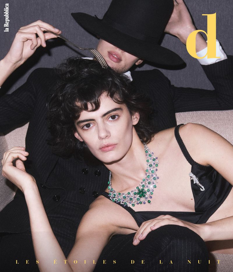 Two fashion models, a woman wearing an emerald necklace and a man in a fedora, pose dramatically for a magazine cover.