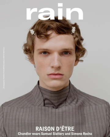Close-up studio shot of a fair-skinned adult male model wearing a gray pinstripe turtleneck, styled with hair clips, with 'rain' logo above.