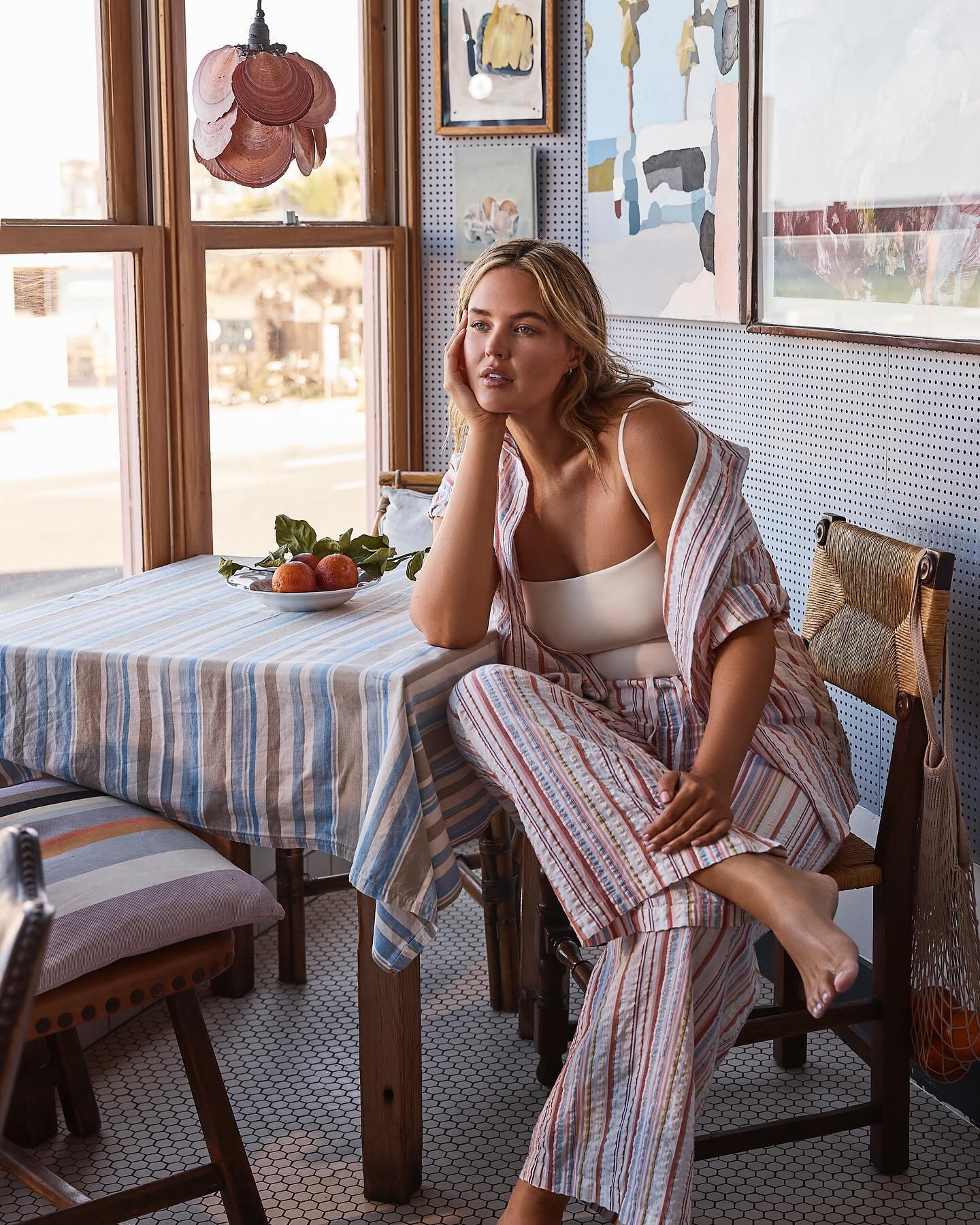 Woman in striped pajamas sitting at a table with oranges near a window, looking contemplative in a well-lit indoor setting.