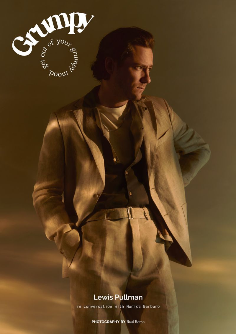 A man in a beige suit poses against a muted gold background; he's captured in a medium shot for 'Grumpy' magazine, in soft, diffused light.