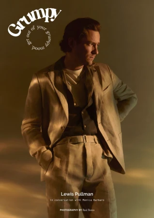 A man in a beige suit poses against a muted gold background; he's captured in a medium shot for 'Grumpy' magazine, in soft, diffused light.