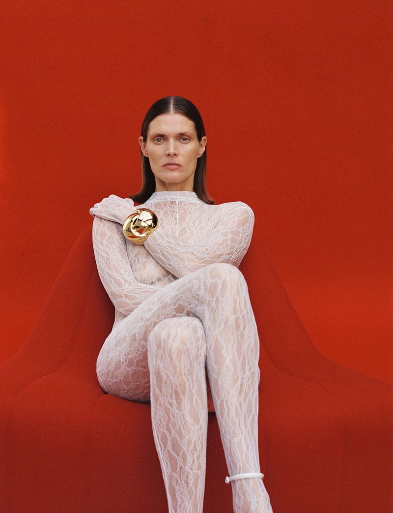 Fashion portrait of a woman with long dark hair wearing a white lace bodysuit and a gold bracelet, posing against a red backdrop.