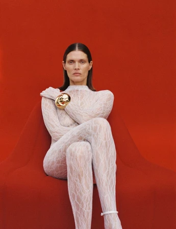 Fashion portrait of a woman with long dark hair wearing a white lace bodysuit and a gold bracelet, posing against a red backdrop.