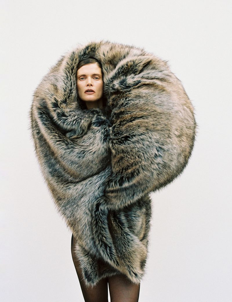 A woman is enveloped in a large, dramatic, grey-brown faux fur wrap against a white background in a fashion-forward portrait.