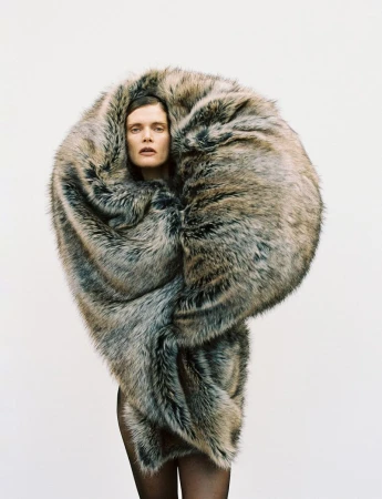 A woman is enveloped in a large, dramatic, grey-brown faux fur wrap against a white background in a fashion-forward portrait.