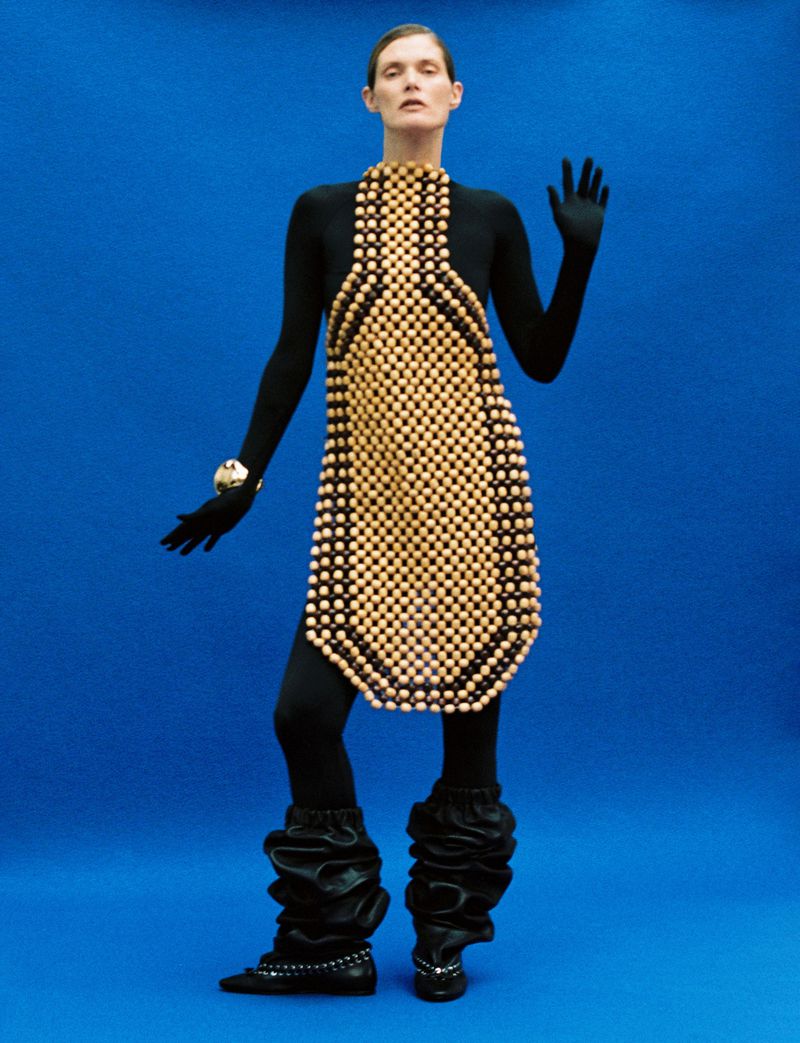 A model poses in a black bodysuit and light brown wooden beaded dress against a solid blue backdrop in a fashion forward shot.