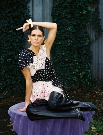 A woman sits on a table outdoors, wearing a polka dot dress, gold jewelry, and black pants, with a green foliage background.