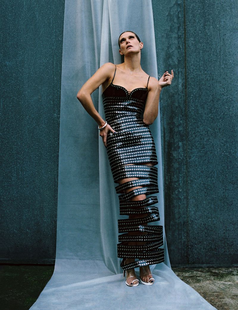 Adult female in a black and silver dress with cutout detail, posing in front of a mottled grey wall and a sheer white curtain.