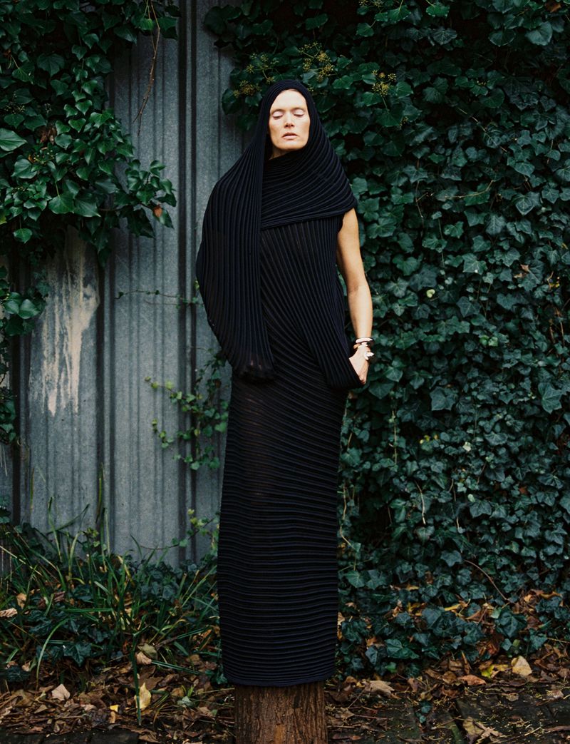 Woman in pleated black dress and hood stands on tree stump in front of green ivy-covered wall, minimalist fashion editorial outdoor.