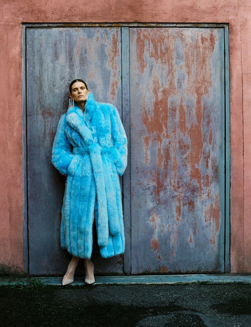 Woman in a long, blue, faux-fur coat poses in front of a textured, pink and gray double-door in an outdoor urban setting.