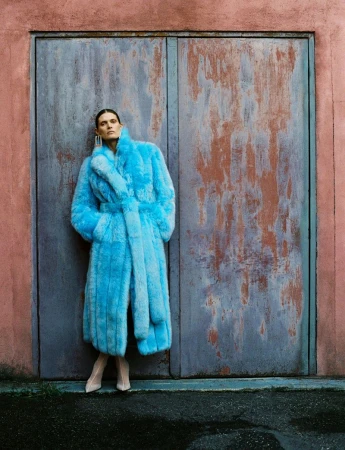 Woman in a long, blue, faux-fur coat poses in front of a textured, pink and gray double-door in an outdoor urban setting.