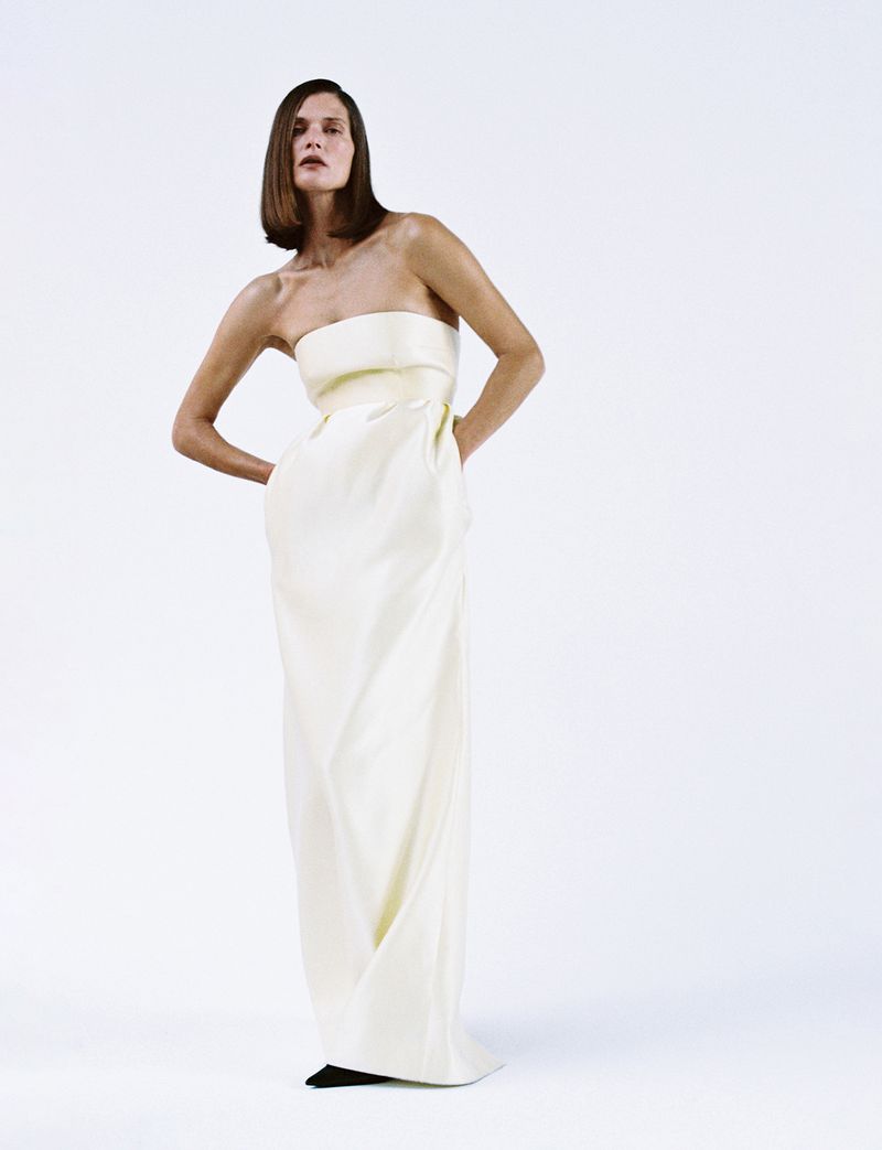 A woman in a white strapless dress poses in a bright studio with her hands on her hips, standing against a clean white backdrop.
