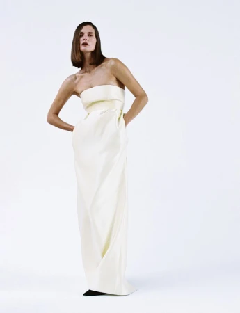 A woman in a white strapless dress poses in a bright studio with her hands on her hips, standing against a clean white backdrop.